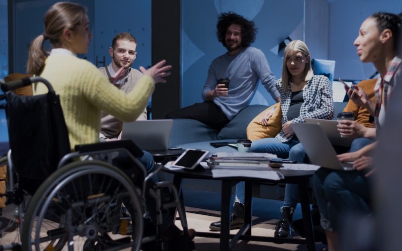 Handicapped young woman with colleagues working in office. She is smiling and passionate about the workflow. Performing in co-working space. Office people working together.